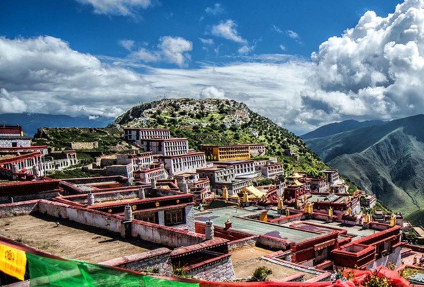 Ganden Monastery, with its red roof and surrounded by clouds in the sky, sits perched atop a hillside with a stunning view of the surrounding landscape.