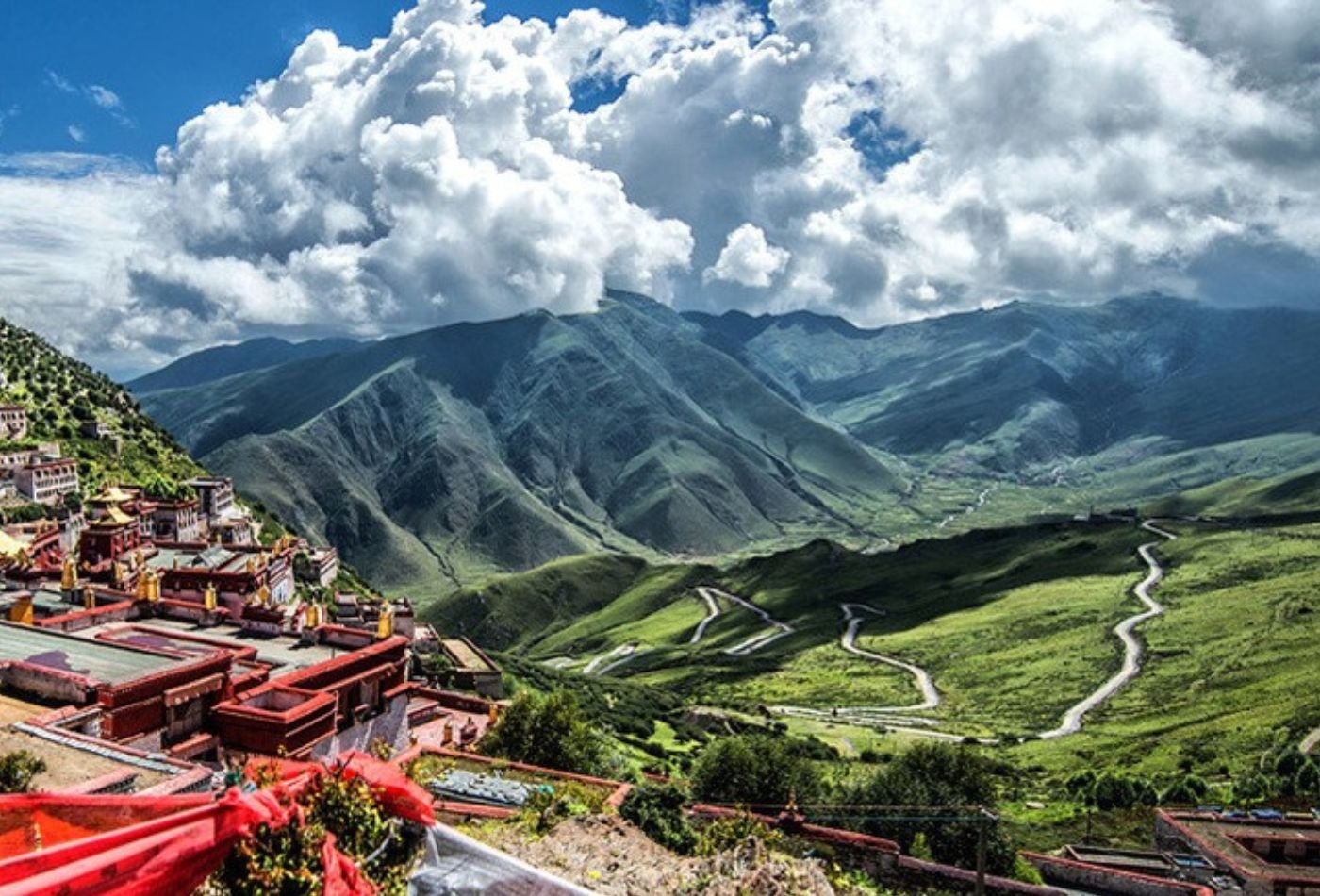A winding road leads to Ganden Monastery, nestled in a valley with clouds in the sky above. The surrounding landscape is lush and green, adding to the beauty of the scene.