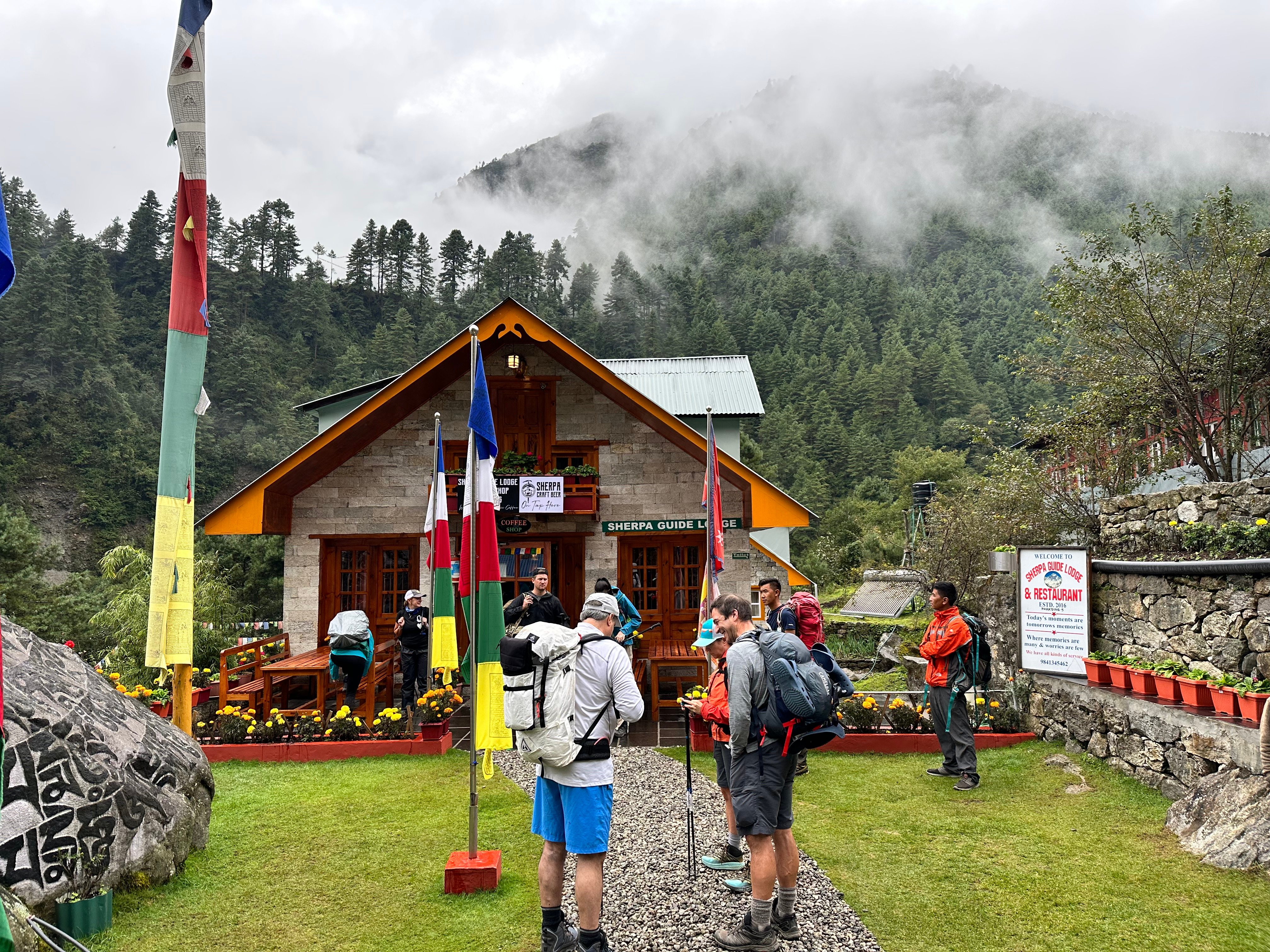 A view of Sherpa Guide Lodge in Phakding, surrounded by lush green trees and vegetation. Several trekkers are seen getting ready for their hike, with backpacks and trekking poles in hand. 