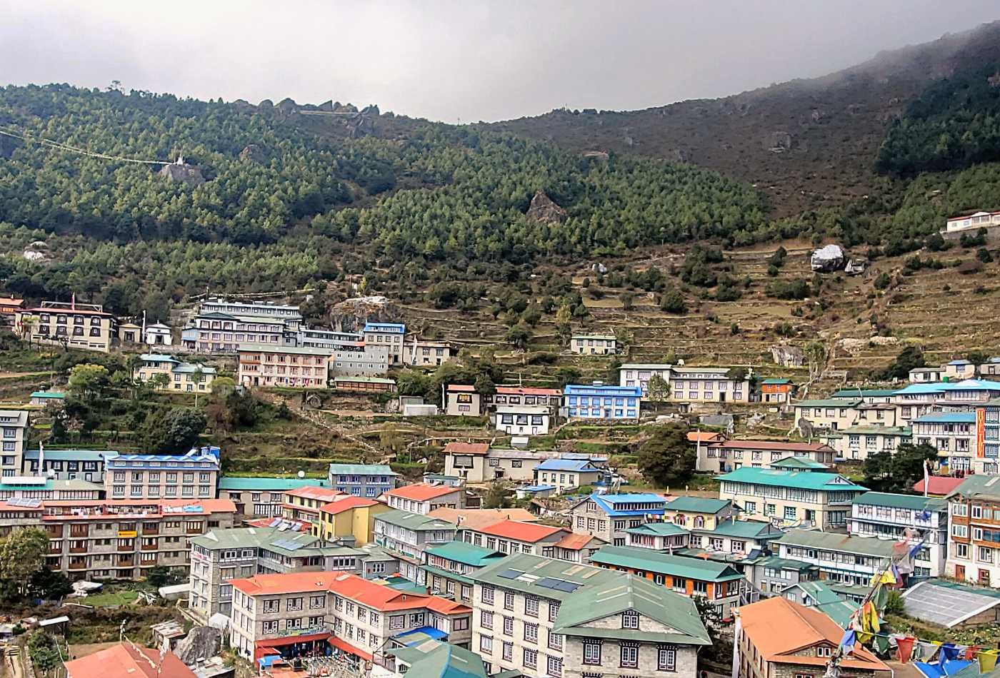 A view of Namche Bazaar, a colorful Himalayas village with a traditional rooftop decorated with prayer flags and surrounded by green hills.