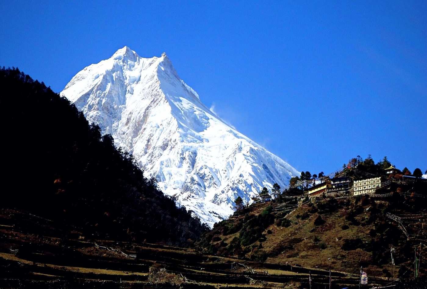 A breathtaking image of the snow-capped summit of Mt. Manaslu, set against a clear blue sky and the picturesque village of Samagaon in the foothill.