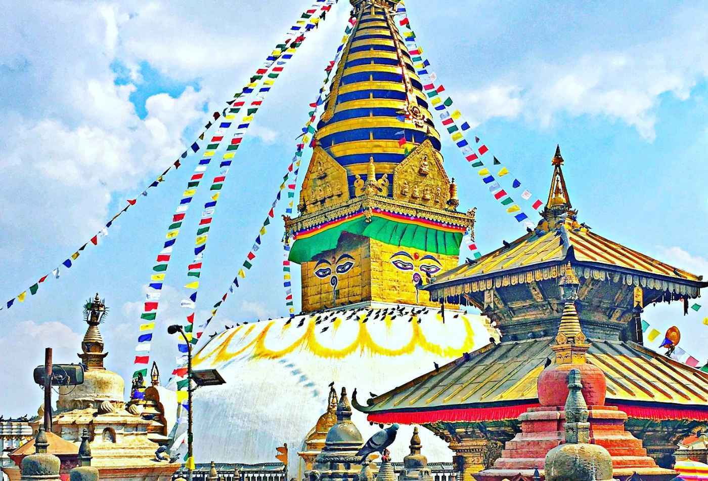 Colorful prayer flags surround the Swayambhunath stupa, and  A smaller temple can be seen standing beside it, with traditional Nepalese architecture.