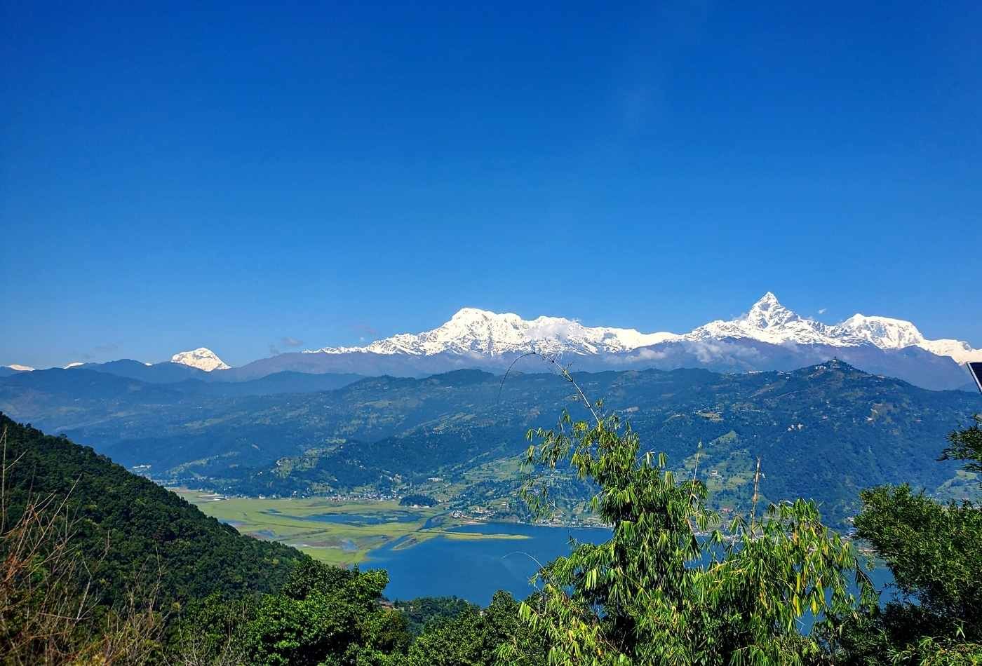 A panoramic view of the Annapurna mountain range, as seen from the Peace Stupa in Pokhara. In the foreground, there is a green valley and a lake.