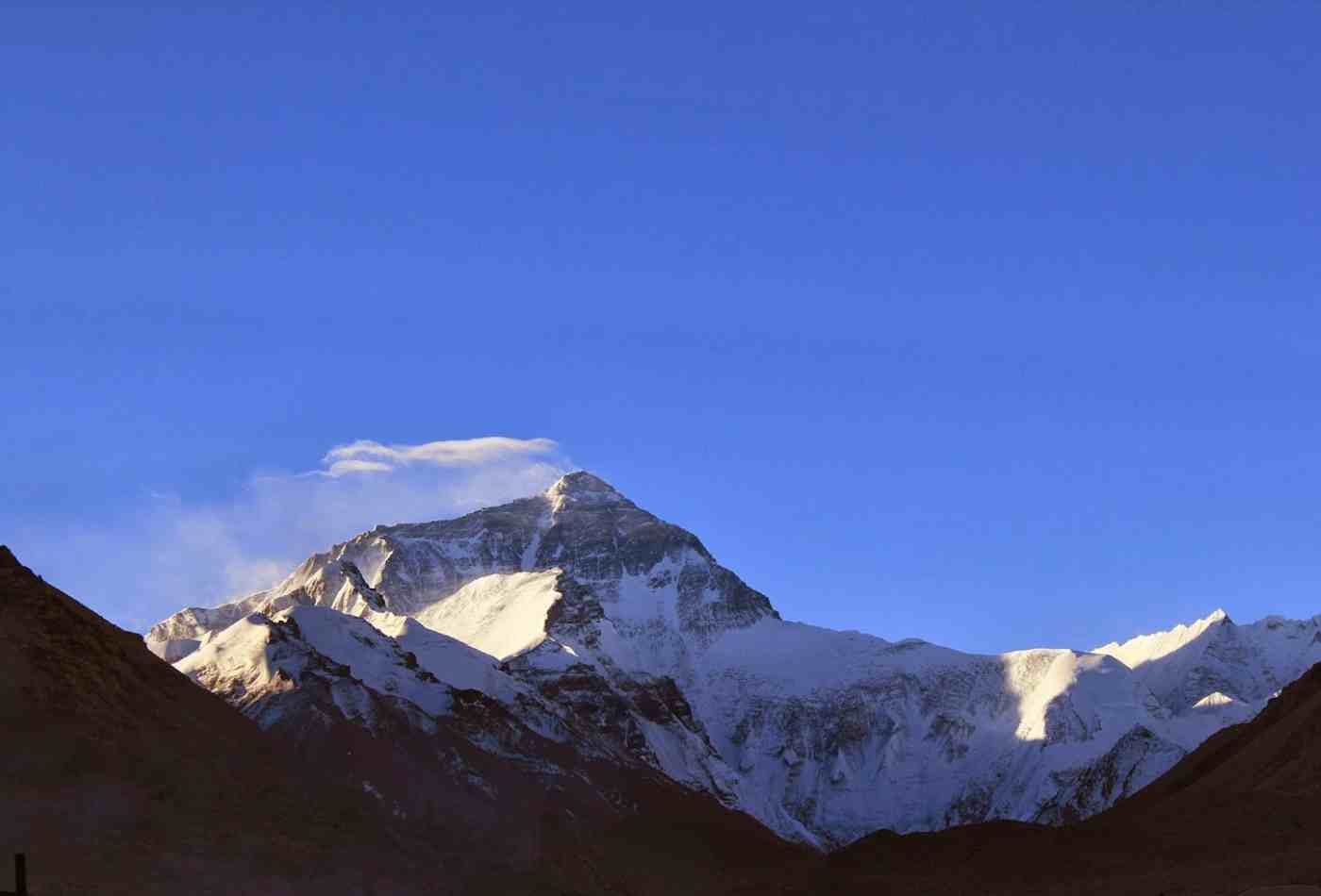 On a clear day, Mt. Everest towering above the clouds, as seen from the Tibet side. The summit and ridges are visible against a bright blue sky.