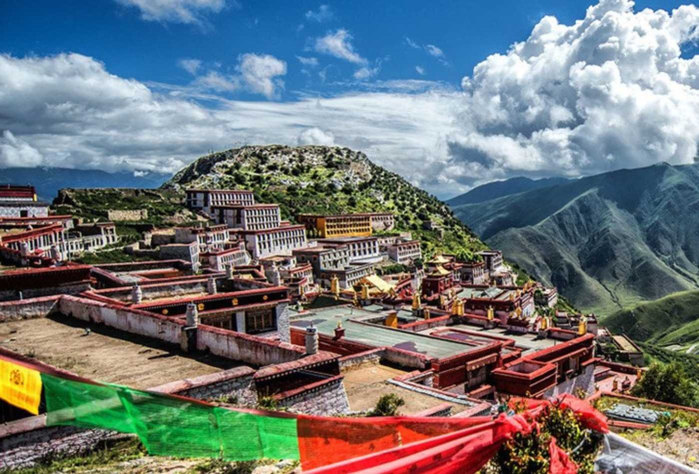 Ganden Monastery, with its red roof and surrounded by clouds in the sky, sits perched atop a hillside with a stunning view of the surrounding landscape.