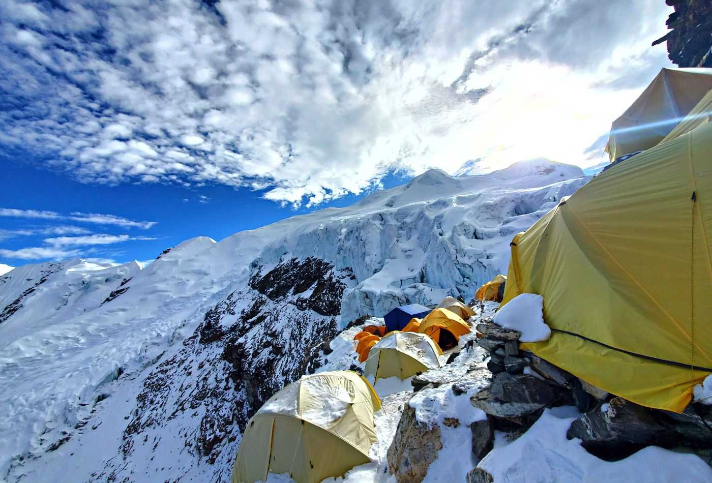 A campsite at Mera High camp with a tent set up on the snowy ground, surrounded by majestic mountains, with white clouds filling the blue sky.