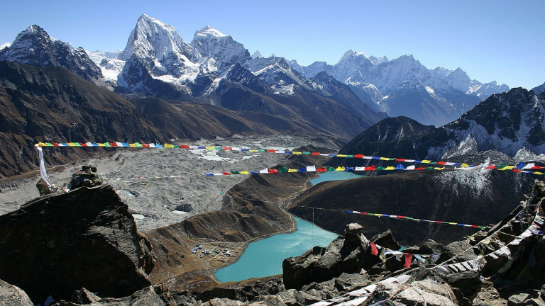 A breathtaking view of Gokyo-Ri with Gokyo Lake in the foreground, surrounded by majestic snow-capped mountains. A line of colorful player flags flutters in the breeze, adding to the beauty of the scene. 
