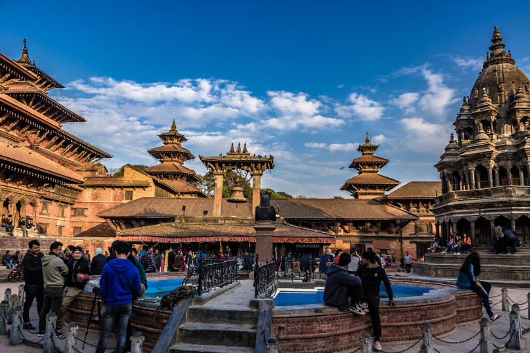 A view of Patan Durbar Square, with a towering temple on the left and a big bell hanging in the middle of the square. The sky is partly cloudy with scattered clouds.